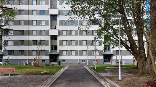 Asuinrakennus piha-alueineen, keskellä kulkee kävelytie ja ympärillä puita, penkkejä ja pyörätelineitä.