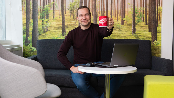 Eevertti Jurvanen istuu sohvalla, hymyilee ja kippistää punaisella kahvimukilla kuvan katsojalle.