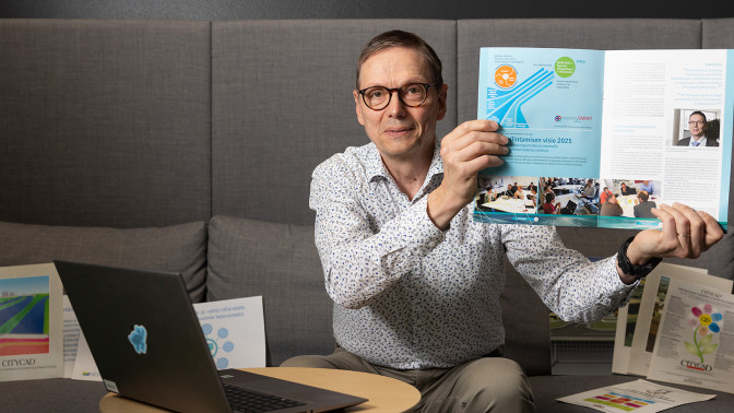 Juha Liukas istuu sohvalla tietokoneen äärellä ja näyttää kameralle uransa alkupuolen paperista esitettä.