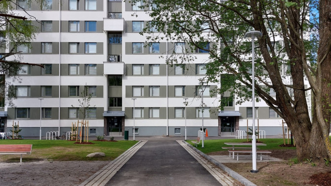 Asuinrakennus piha-alueineen, keskellä kulkee kävelytie ja ympärillä puita, penkkejä ja pyörätelineitä.