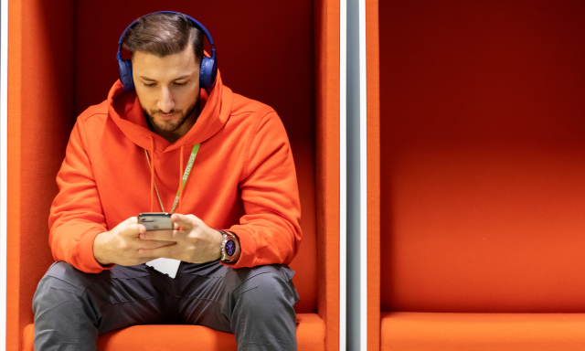 Henkilö istuu yksin oranssissa puhelinkopissa ja tutkii puhelintaan.