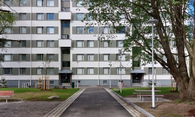 Asuinrakennus piha-alueineen, keskellä kulkee kävelytie ja ympärillä puita, penkkejä ja pyörätelineitä.