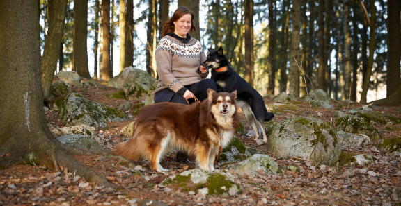 Aino Karilas istuu metsässä kivellä ja kaksi koiraa seisoo hänen jaloissaan.