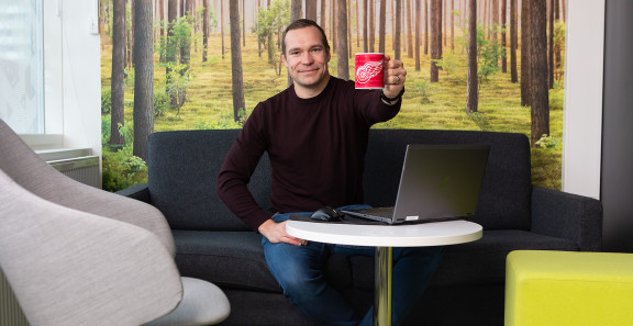 Eevertti Jurvanen istuu sohvalla, hymyilee ja kippistää punaisella kahvimukilla kuvan katsojalle.