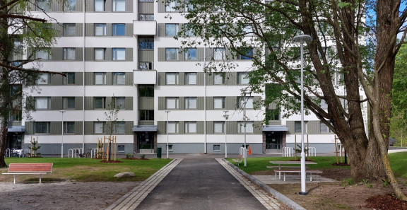 Asuinrakennus piha-alueineen, keskellä kulkee kävelytie ja ympärillä puita, penkkejä ja pyörätelineitä.