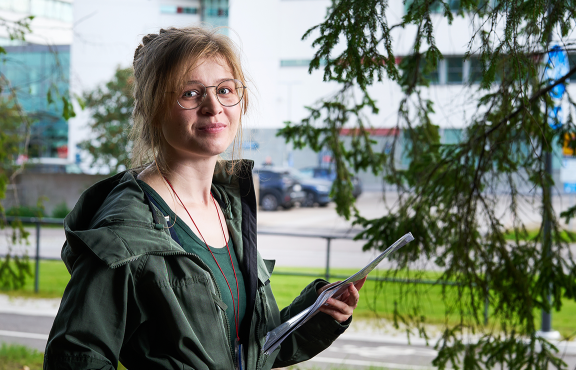 Heli Vainio seisoo muistiinpanokansion kanssa metsänreunassa kaupunkiympäristössä. 