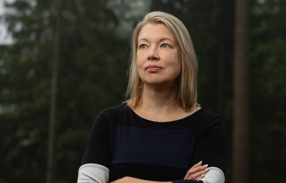 Nora Sillanpää seisoo metsän edustalla luoden rauhallisen katseen kameran ohi.