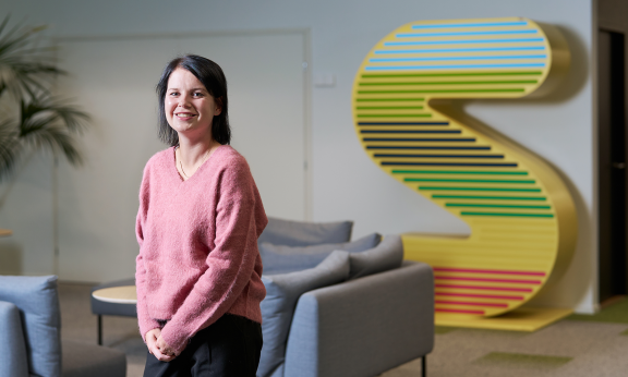 Emma Liljeström seisoo toimiston aulassa ja hymyilee kameralle.