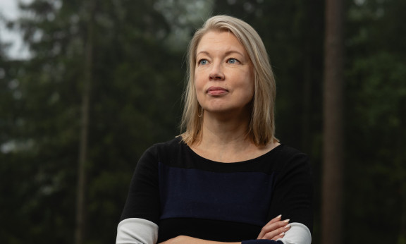 Nora Sillanpää seisoo metsän edustalla luoden rauhallisen katseen kameran ohi.