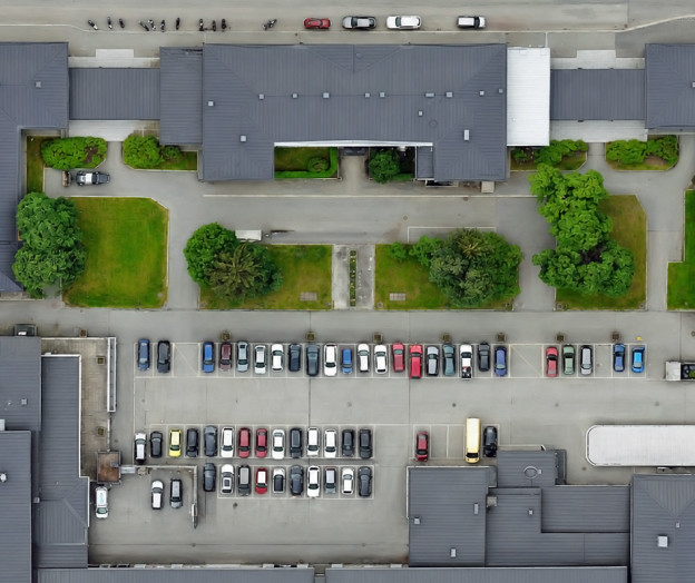 Ilmakuva modernista rakennuskokonaisuudesta, jossa on harmaakattoisia rakennuksia, sisäpihoja, vihreitä istutusalueita ja pysäköintialue autoineen.