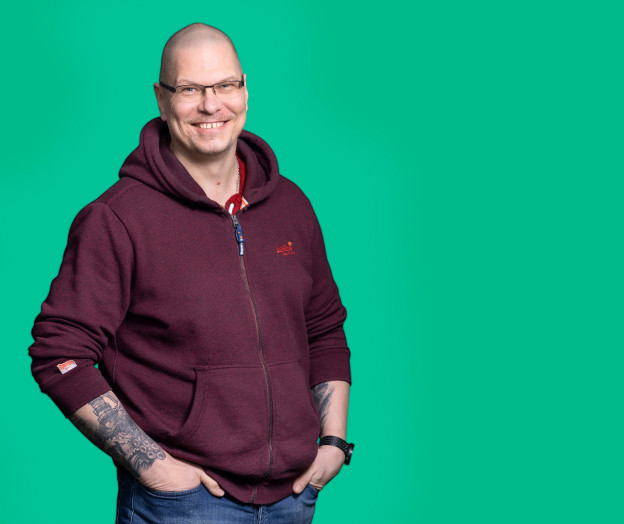 Sitowiseläinen seisoo rennosti kädet taskuissa viininpunaisessa hupparissa vihreää taustaa vasten.