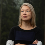 Nora Sillanpää seisoo metsän edustalla luoden rauhallisen katseen kameran ohi.